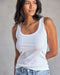 A woman wearing a white ribbed tank top and blue jeans.