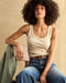 A woman sitting wearing a beige ribbed tank top and blue jeans.