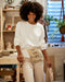 A woman standing in a craft room wearing a white long-sleeved t-shirt with side slits, beige pants, and carrying a beige bag with a text pattern.